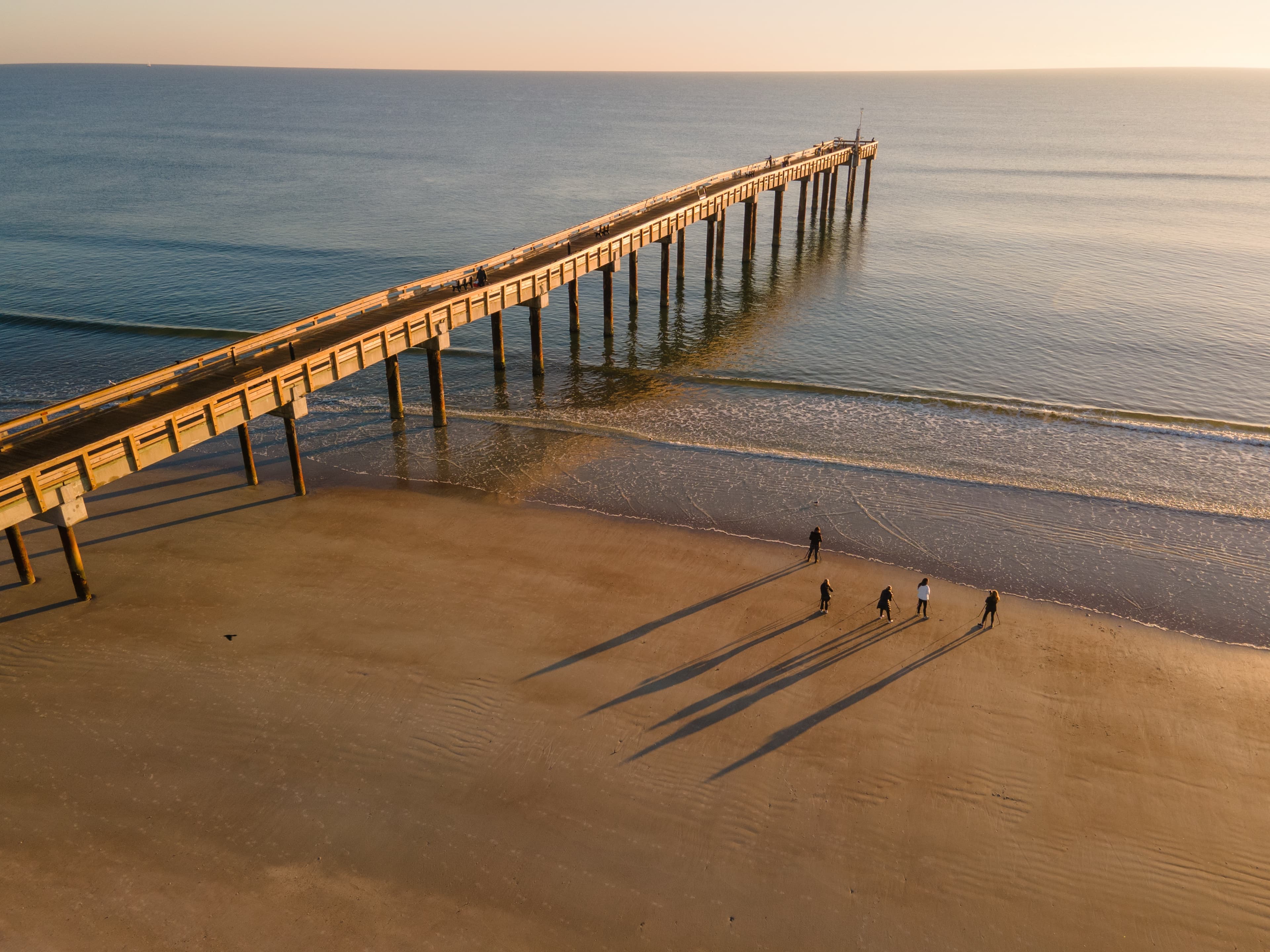 Vista aérea de un grupo con trípodes sobre la arena al atardecer, junto a un muelle largo que entra en el mar.